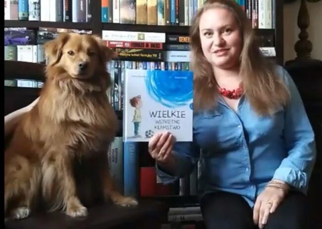 uśmiechnięta bibliotekarka, obok pies siedzi na krześle; bibliotekarka pokazuje książkę pt. Wielkie wstrętne kłamstwo