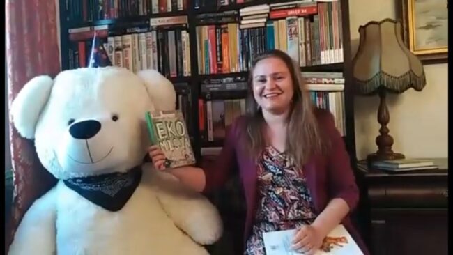 uśmiechnięta bibliotekarka, obok niej wielka maskotka - miś, w tle regał wypełniony książkami, bibliotekarka pokazuje książkę pt. Ekomisja