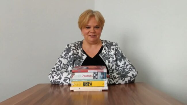 uśmiechnięta bibliotekarka, przed nią na stoliku stosik książek omawianych podczas tego przeglądu nowości