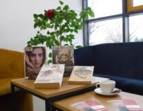zdjęcie przedstawia książki wyeksponowane na stoliku, dotyczące tematu spotkania; obok książek stoi filiżanka, na spodku jest też ciastko - delicja; wokół stolika sofa i fotele, kwiaty doniczkowe