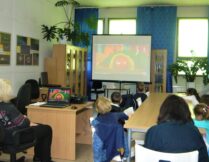dzieci siedzą przy stolikach i oglądają bajkę o jesieni, wyświetlaną na ekranie projektora
