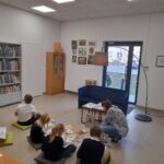 dzieci siedzą na poduszkach na podłodze biblioteki i słuchają książki czytanej przez bibliotekarkę