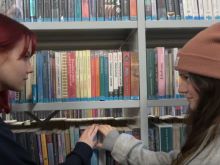 uczestniczki projektu sięgają po tę samą książkę na regale w bibliotece  i patrzą na siebie uśmiechnięte