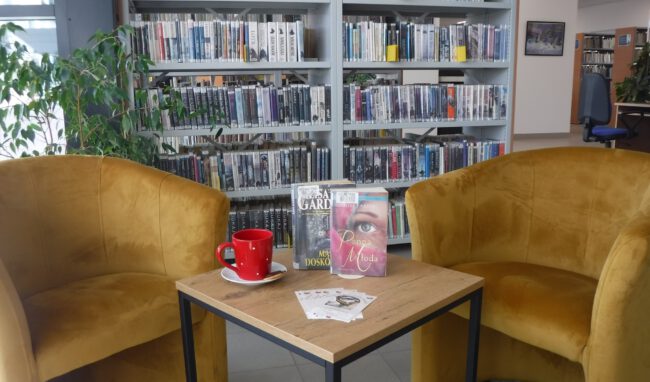 na bibliotecznym stoliku są książki omawiane poczas spotkania i kubek; obok fotele, w tle regały biblioteczne