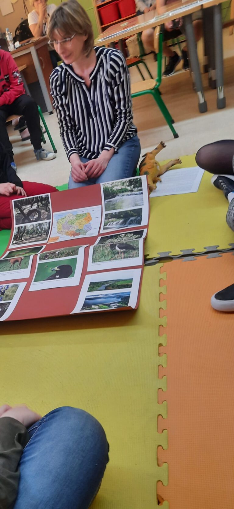 Pani bibliotekarka prezentuje uczniom planszę przedstawiająca najdziksze zakątki Polski i świata.