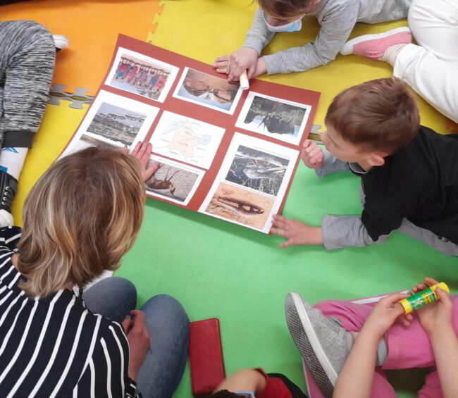 Pani bibliotekarka prezentuje uczniom planszę przedstawiająca najdziksze zakątki Polski i świata.