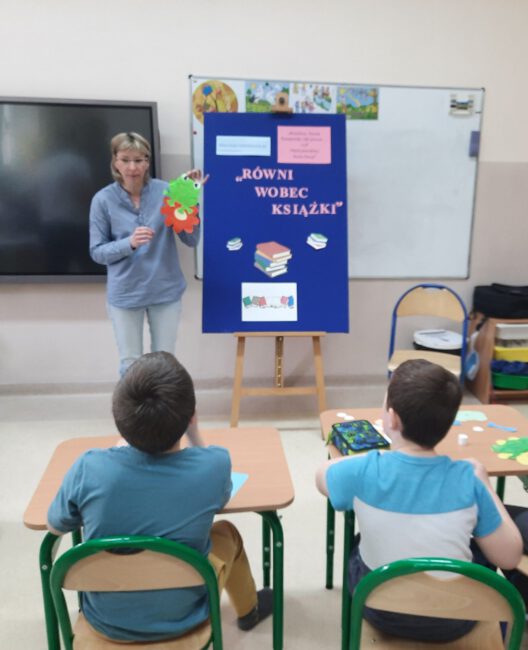 Pani bibliotekarka prezentuje uczniom plansze, na której znajduje się informacja o temacie warsztatów oraz pokazuje prototyp pracy plastycznej, którą uczniowie wykonają tj. żabkę.