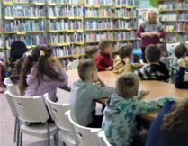 dzieci siedzą przy stolikach i słuchają bibliotekarki