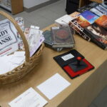 wiklinowy koszyk, a w nim zakładki do książek i logotyp wydarzenia; obok pamiątkowe pieczątki i książki - nagrody w quizie