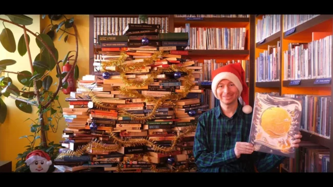 bibliotekarz czyta w czapce mikołaja fragment książki na tle choinki z książek