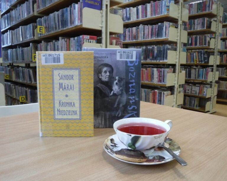 filiżanka z herbatą, obok książki na stoliku, w tle regały z książkami