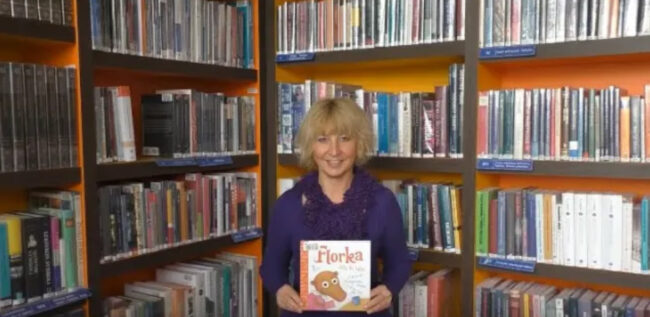 uśmiechnięta bibliotekarka trzyma w dłoniach książkę Florka. Listy do babci, za bibliotekarką regał wypełniony książkami