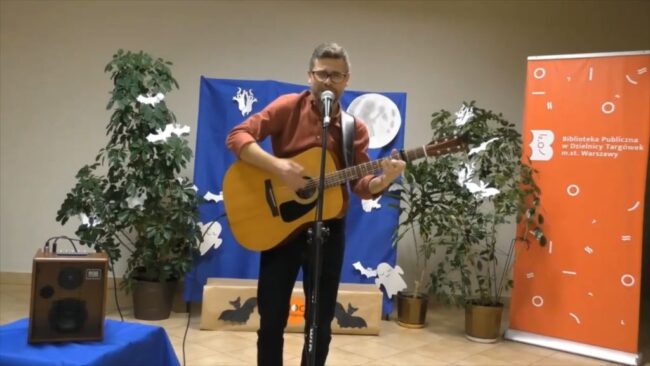 Jacek Gutry stoi przy mikrofonie, śpiewa i gra na gitarze; po bokach duże rośliny doniczkowe