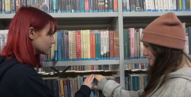 dwie licealistki wpatrzone w siebie, dotykające swoich dłoni; w tle regał wypełniony książkami