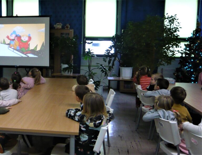 dzieci siedzą przy stolikach i oglądają prezentację multimedialną w bibliotece