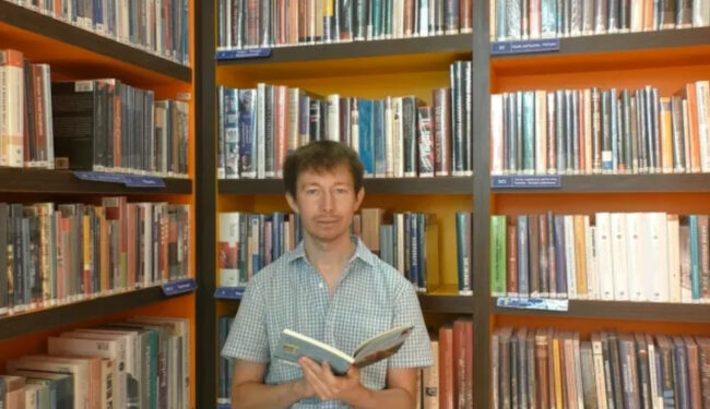 bibliotekarz trzyma książkę; w tle regał wypełniony książkami