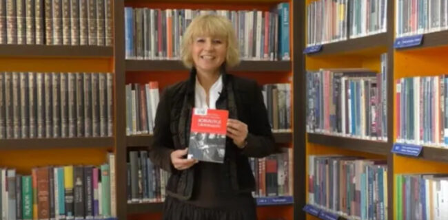 uśmiechnięta bibliotekarka trzyma w dłoniach książkę Korepetycje z niepodległości, za nią regał wypełniony książkami