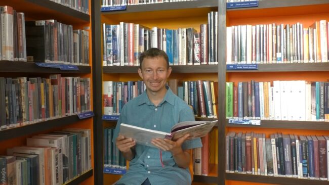 uśmiechnięty bibliotekarz czyta książkę Maleńkie królestwo królewny Aurelki, za bibliotekarzem regał wypełniony książkami
