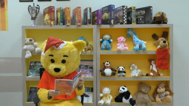 maskotka biblioteki - miś - siedzi na tle maskotek i czyta książkę; na głowie ma czapkę mikołaja