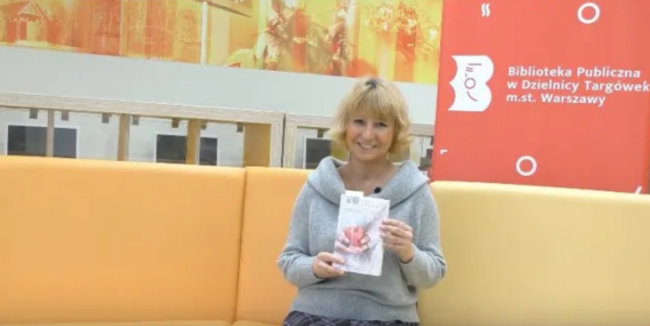 uśmiechnięta bibliotekarka trzyma w dłoniach książkę pod tytułem Niemłodość, siedząc na sofie w czytelni naukowej nr 1