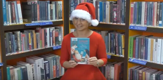 bibliotekarka w czapce mikołaja i czerwonej sukience trzyma w dłoniach książkę Pamiętnik świętego Mikołaja