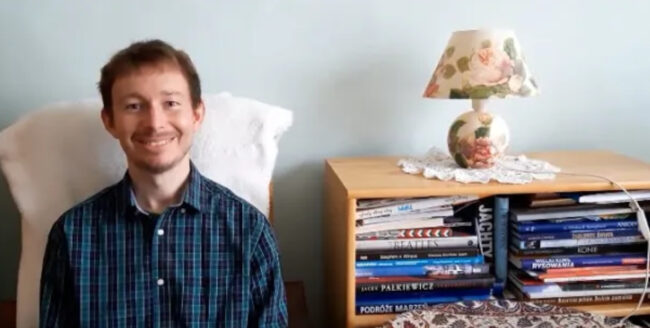 uśmiechnięty bibliotekarz siedzi na hotelu, obok regał wypełniony książkami, na nim lampka