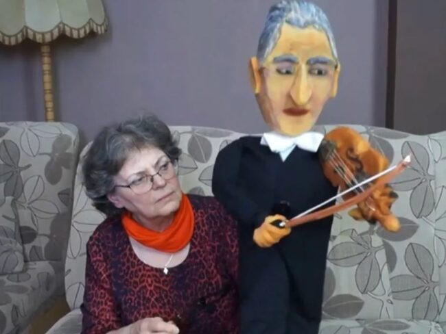 kobieta siedzi na sofie, trzyma dużą lalkę - mężczyznę grającego na skrzypcach