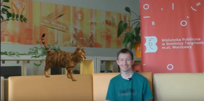 uśmiechnięty bibliotekarz siedzi na sofie, obok niego animacja - kot stojący na oparciu sofy