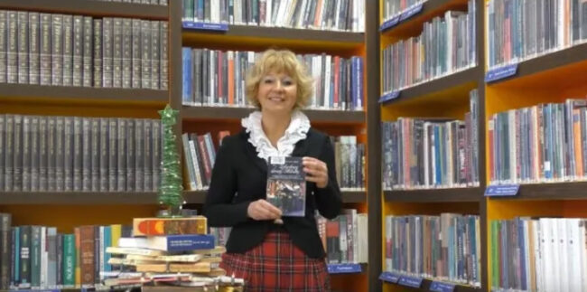 uśmiechnięta bibliotekarka trzyma w dłoniach książkę Zakochany święty Mikołaj, stoi na tle regału bibliotecznego, przy choince