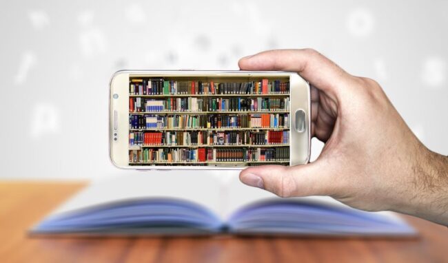 dłoń trzymająca smartfon, na ekranie regał wypełniony książkami, w tle za smartfonem na blacie biurka leży otwarta książka