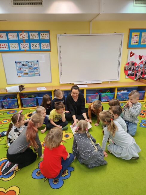 dzieci słuchają książki czytanej przez bibliotekarkę; wszyscy siedzą na podłodze