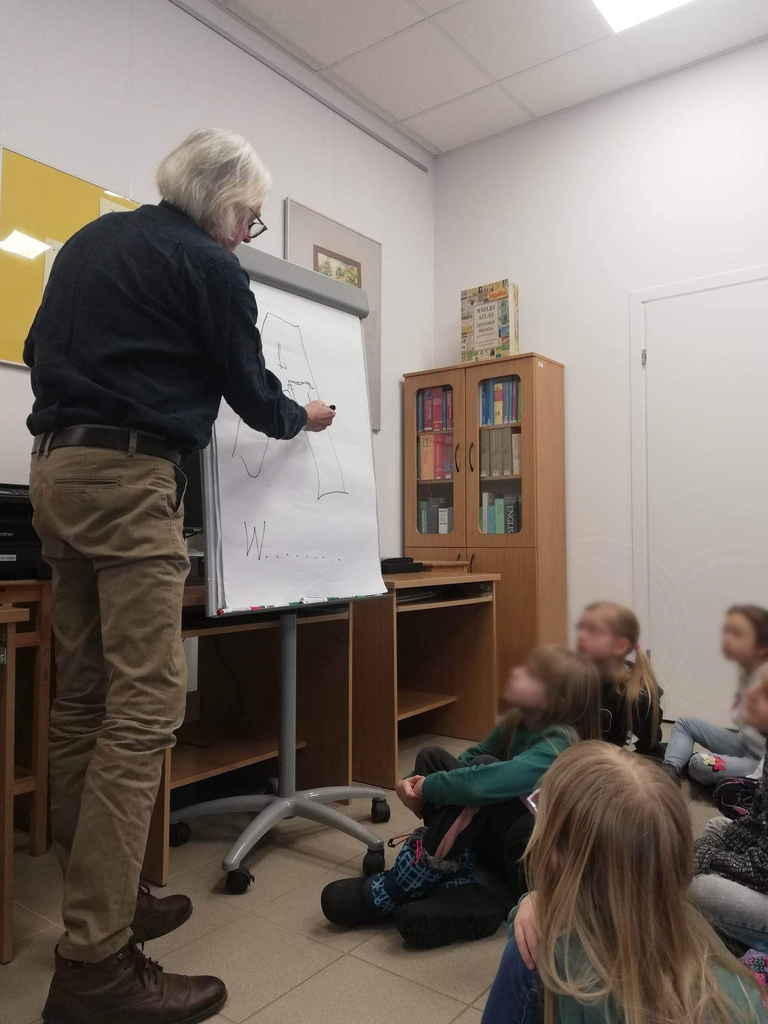 Marek Samselski rysuje rebusy na kartce papieru; dzieci siedzą na podłodze i obserwują