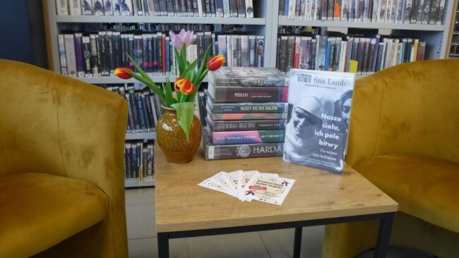Na tle regału bibliotecznego stolik, na którym znajdują się książki, tulipany w wazonie, ulotki promujące spotkanie DKK.