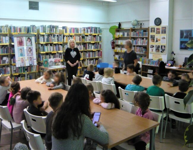 dzieci słuchają pani bibliotekarki opowiadającej o emocjach; w tle regał z książkami