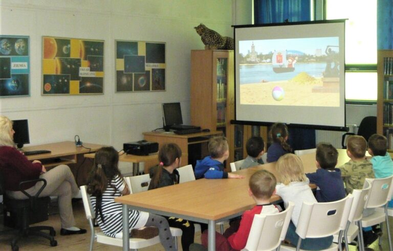 dzieci siedzą przy stolikach i oglądają na ekranie projektora prezentację multimedialną