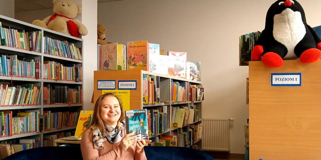 uśmiechnięta bibliotekarka pokazuje nowości książkowe dla dzieci i młodzieży; w tle regały, na których są duże maskotki krecika i misia