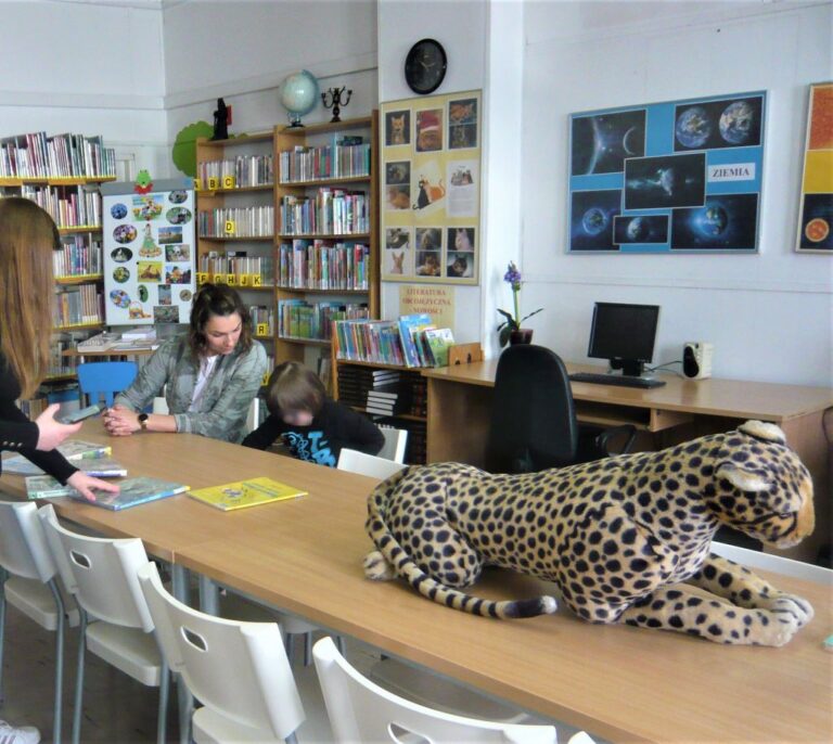 dziewczynka wspólnie z bibliotekarką przegląda książkę, obok na stoliku duża maskotka geparda