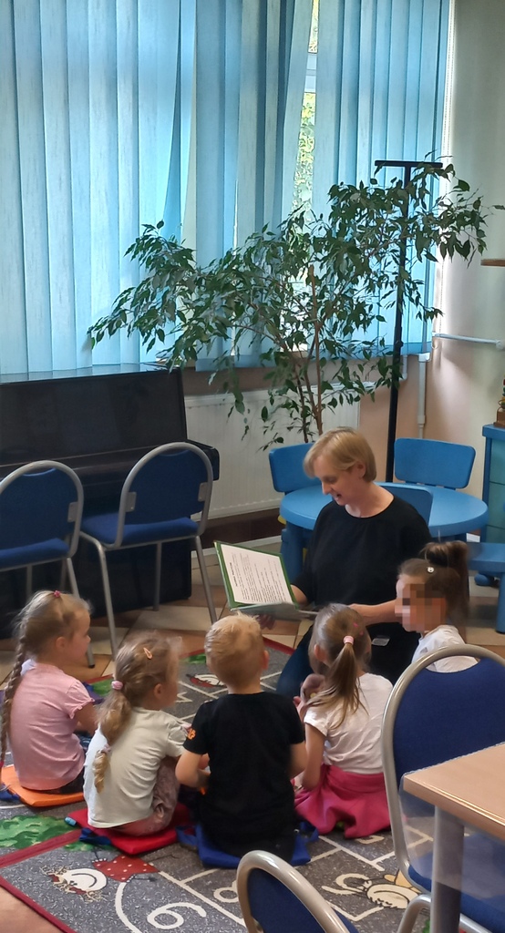 bibliotekarka czyta książkę dzieciom siedzącym wokół niej; wszyscy siedzą na podłodze