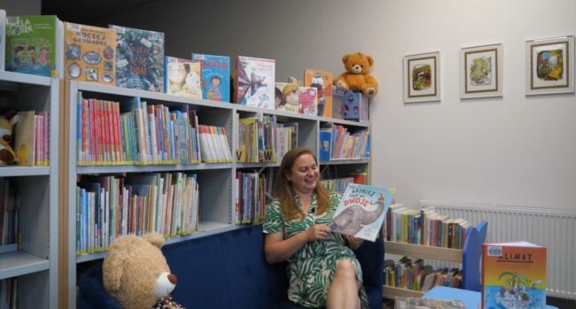 uśmiechnięta bibliotekarka opowiada o polecanych nowościach książkowych dla dzieci i młodzieży; omawia m.in. trzymaną w rękach książkę „Fajniej jest we dwoje" autorstwa Michelle Courtney