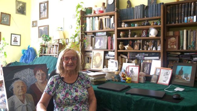 Hanna Budzisz siedzi przy stole; w tle regał z książkami i pamiątkami po Janinie Englert