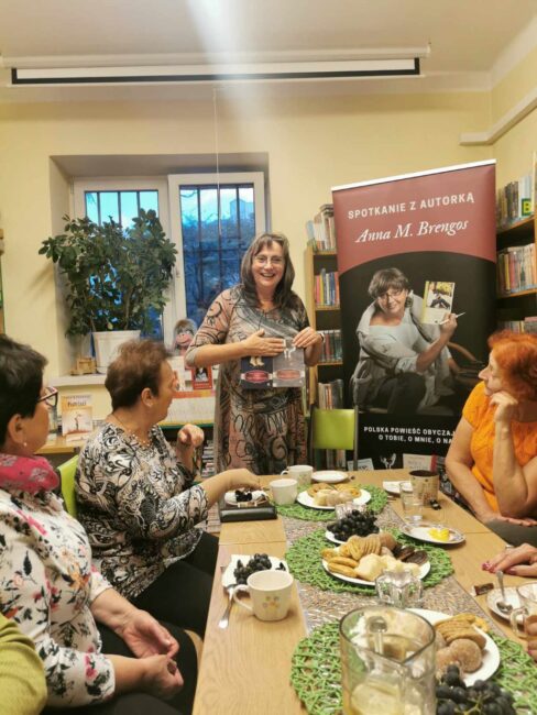 spotkanie autorskie z Anną M. Brengos; uczestniczki siedzą wokół stołu zastawionego poczęstunkiem (ciastka, herbata)