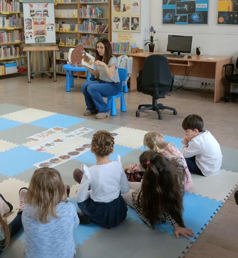 grupa dzieci siedzi na macie w bibliotece i słucha nauczycielki Early Stage