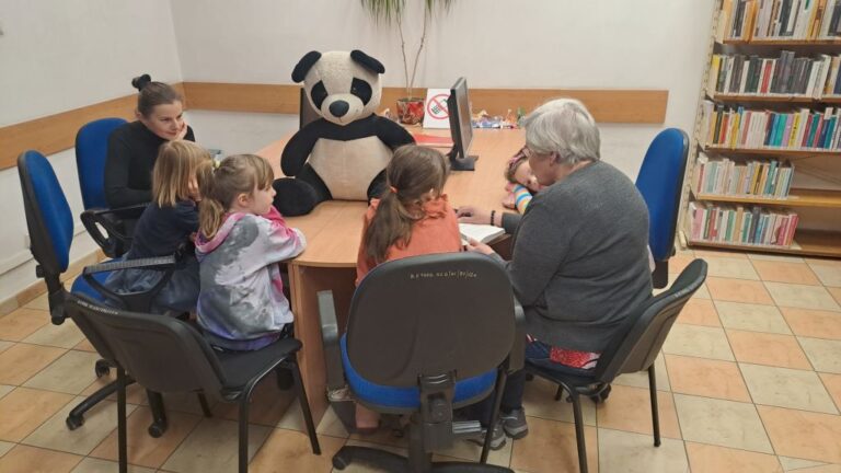 dzieci siedzą przy stoliku razem z dużą maskotką pandy i wolontariuszką, która czyta książkę