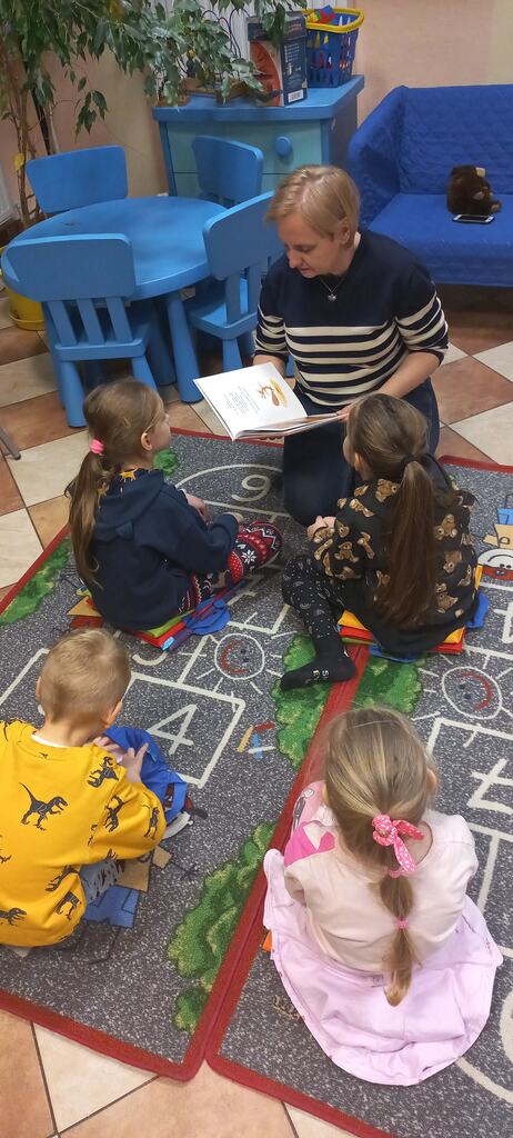 dzieci siedzą na dywanie w bibliotece i słuchają książki czytanej przez bibliotekarkę