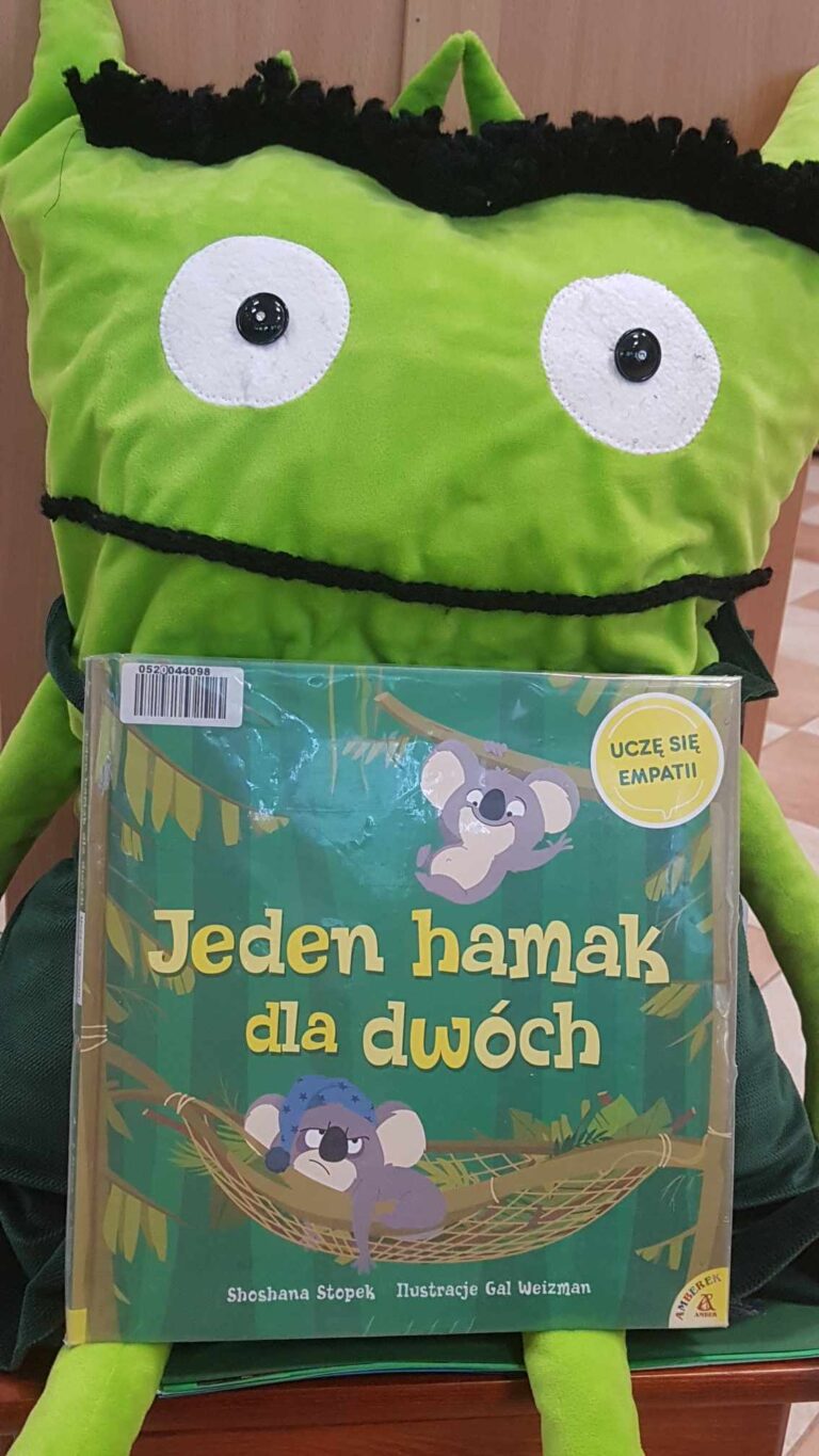 uśmiechnięta maskotka trzyma w łapkach książkę Jeden hamak dla dwóch