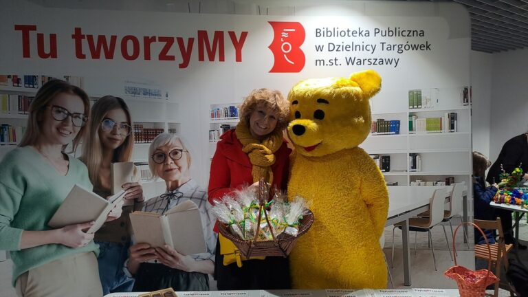 duża maskotka misia trzyma koszyk z upominkami, obok uśmiechnięta animatorka kultury w bibliotece publicznej na Targówku