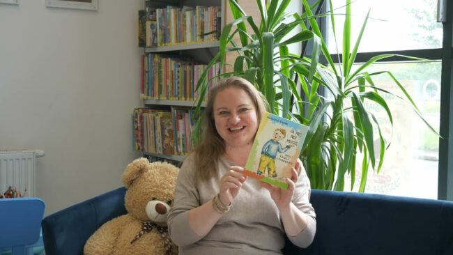 uśmiechnięta bibliotekarka prezentuje nowości wydawnicze, siedzi na sofie, obok niej duża maskotka misia, w tle regał z książkami i duży kwiat doniczkowy