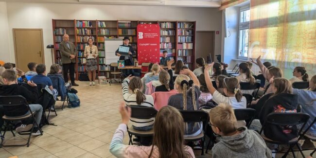 spotkanie autorskie z Wojciechem Widłakiem; na sali kilkadziesiąt osób, głównie uczniowie IV klasy szkoły podstawowej; dyrekcja biblioteki wita gości; Wojciech Widłak trzyma laptop z wyświetlonymi na ekranie uczestnikami spotkania online