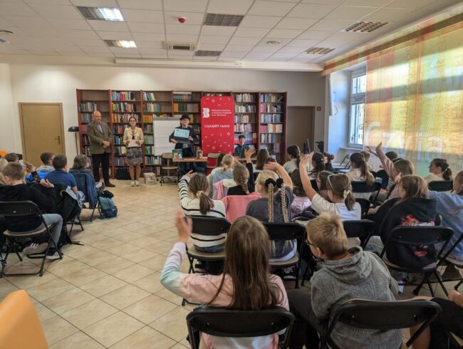 spotkanie autorskie z Wojciechem Widłakiem; na sali kilkadziesiąt osób, głównie uczniowie IV klasy szkoły podstawowej; dyrekcja biblioteki wita gości; Wojciech Widłak trzyma laptop z wyświetlonymi na ekranie uczestnikami spotkania online