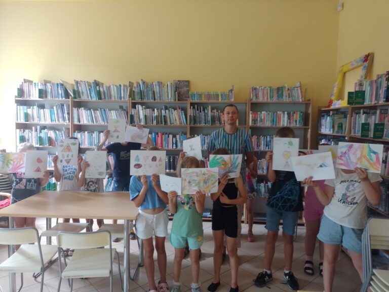 dzieci pokazują narysowane przez siebie obrazki; w tle regały z książkami